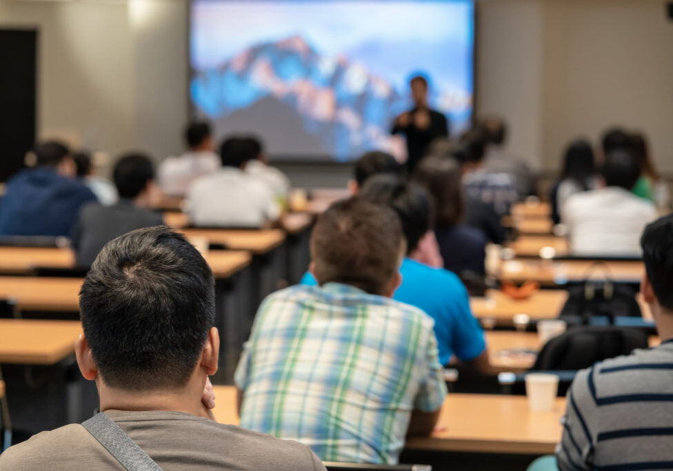 Speaker on the stage in front of the room with Rear view of Audience in put hand up acton for answer the question in the meeting or seminar meeting, business and education concept