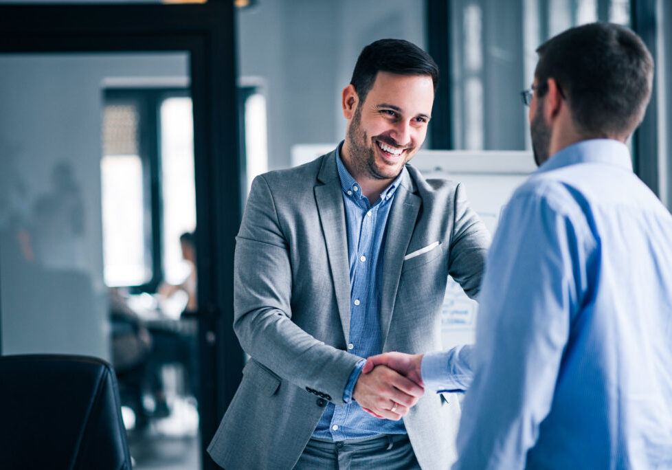 Two smiling businessmen shaking hands while standing in an office.